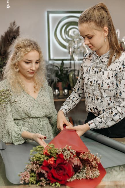Two women are in a floral shop, with one woman, wearing a patterned blouse, carefully wrapping a vibrant bouquet of red roses, pink baby's breath, and greenery in red floral wrapping paper. The other 