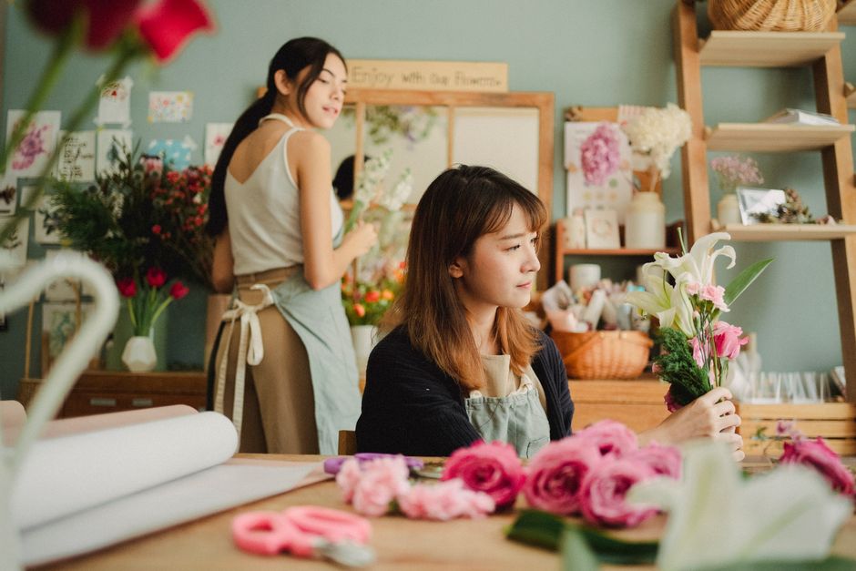 Two women working in a floral shop, Bellingham Flowers, arranging and handling fresh flower bouquets. The woman in the foreground, with shoulder-length brown hair, is holding a floral arrangement comp