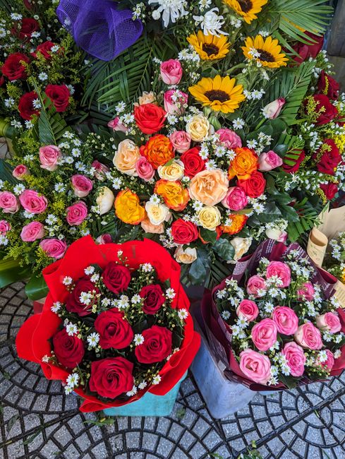 A vibrant display of fresh floral arrangements on a cobblestone surface, featuring a large bouquet of multicolored roses including pink, red, peach, and orange, accented with small white filler flower